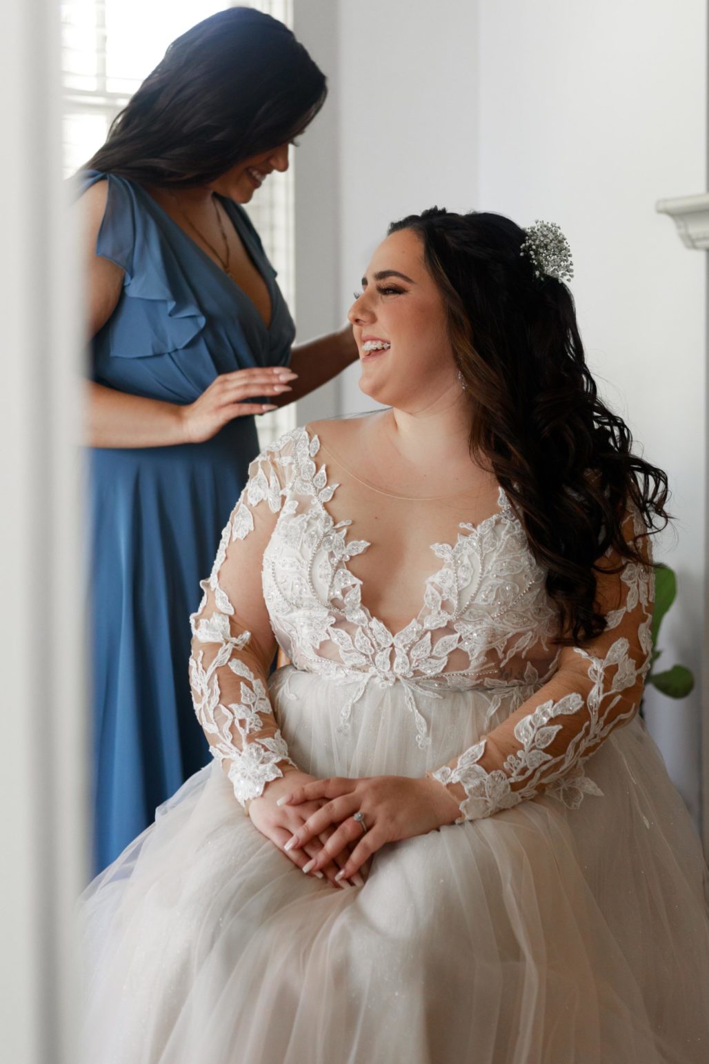 Bride to be is getting ready with her bridesmaid sitting joyfully near a window.