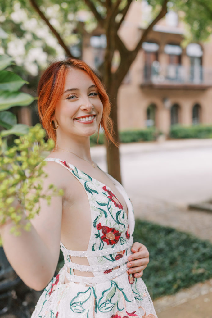   a close-up of a young female College student smiling outside enjoying the outdoor wearing a beautiful white long prom dress that has red and green flowers on it. 