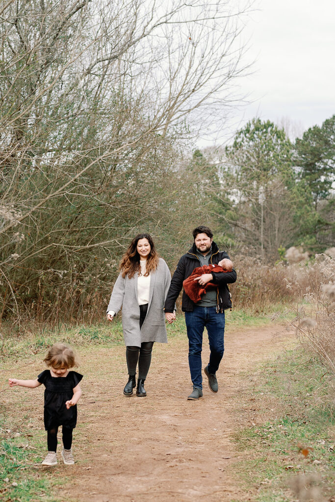 A joyful family; mom, dad, girl toddler and newborn baby boy walking towards a Zebulon GA photography taking their family portraits outside park